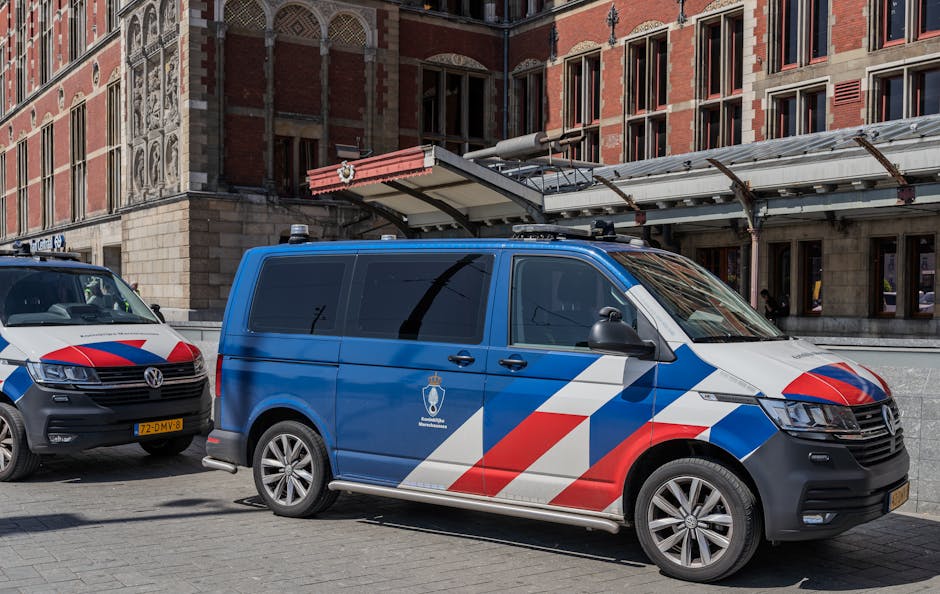 Two police vehicles are parked outside a building with red brick and stone façade, in an urban setting. The vehicle in the foreground is a blue Volkswagen van with a white and red diagonal stripe pat