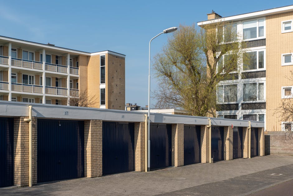 A row of six garage units with dark blue, vertically ribbed metal doors set within low brick walls, with each door numbered sequentially from 67 to 72, situated below a concrete overhang. The pavement in front of the garages is made of grey bricks, and a tall, curved streetlamp stands between garage 68 and 69, casting light over the area. Behind the garages, there are multi-storey residential buildings with balconies, windows, and a beige brick exterior, and a sizable tree with green and yellow foliage. The scene is illuminated by natural daylight, indicating a clear weather condition. Although there are no visible moving activities or objects related to house removals, the setting hints at an urban environment relevant to relocating or transporting household items within or around residential areas, which might be associated with professional moving services such as those offered by Man and Van Holland Park.