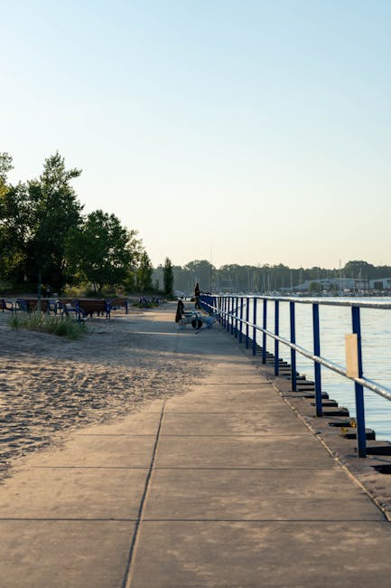 A concrete pathway along a waterfront during daytime, with a blue metal railing on the right side and trees on the left. Several small boats are docked in the distance across the water, and a few people are visible further along the pathway, some seated and others walking. The scene is well-lit with natural daylight, and the pathway appears clean and unobstructed, suitable for walking and transportation. This setting could be part of a home relocation or moving logistics process, as a moving company like Man and Van Holland Park might use such an area for overseeing furniture transport or loading processes for house removals in the Holland Park area. The image captures the outdoor environment where moving activities might take place during a residential move near the water, with a focus on smooth access for loading and unloading items as part of professional relocation services.