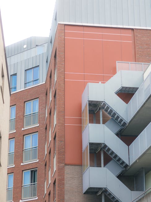 The image depicts the exterior of a multi-storey residential or commercial building with a prominent exposed staircase structure on the right side. The staircase is made of metal with a light grey finish, featuring landings and outdoor stairs that connect multiple levels. Adjacent to the staircase, the building's facade combines red brickwork with sections of grey metal cladding near the roof. Several windows with blue-tinted glass and small metal railings are visible on the brick sections. The scene is captured in natural daylight, with a clear view of the building's modern architectural design. This setting illustrates the type of urban environment where professional house removals or furniture transport services, such as those provided by Man and Van Holland Park, may operate, especially when navigating building access points during a home relocation or similar moving project.