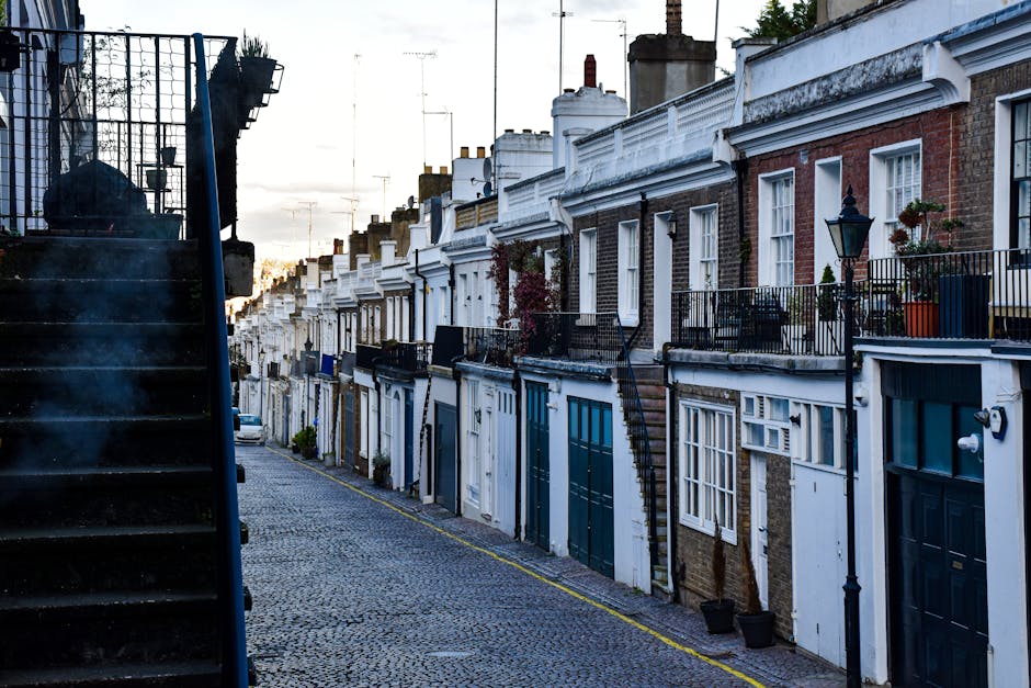 A narrow cobbled street in Holland Park lined with rows of terraced houses featuring brick and white-painted facades, decorative railings, and small front gardens with potted plants. The image captures an outdoor scene during daylight with soft natural lighting, showing a staircase on the left side of the image leading up to an entrance, which may pose access challenges for home relocation or furniture transport. The street appears to have limited space for large vehicle parking or direct load-in, highlighting the need for specialist removals solutions such as those provided by Man and Van Holland Park. The image emphasizes the importance of careful planning in packing and moving tasks when working within constrained environments like tight stair access and narrow alleyways, typical of properties in the Holland Park area. The scene illustrates the residential environment where efficient load handling and home relocation logistics are essential for smooth removals, with minor details like a few potted plants, street lamps, and the subtle glow of evening light enhancing the setting's character.