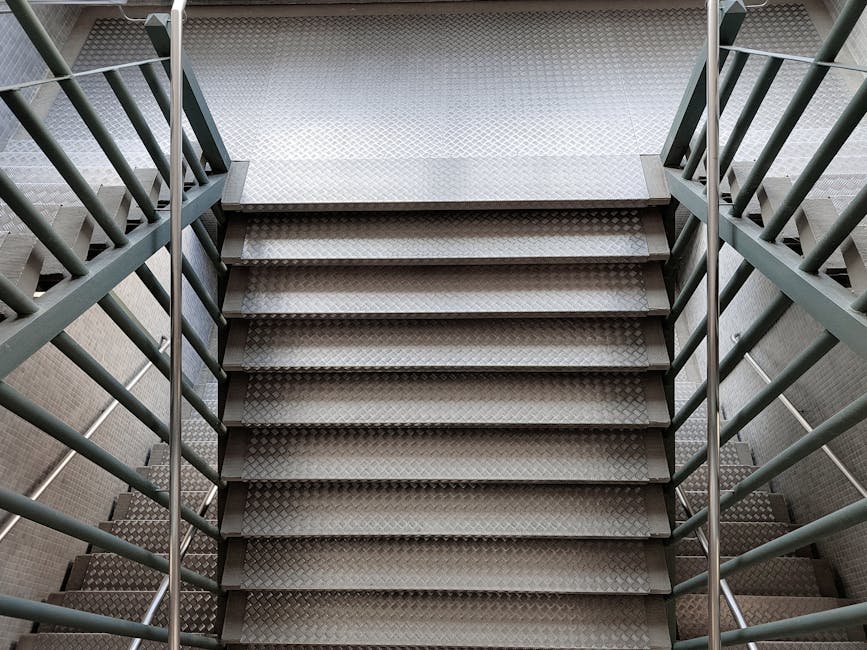The image depicts the exterior of a multi-storey residential or commercial building with a prominent exposed staircase structure on the right side. The staircase is made of metal with a light grey finish, featuring landings and outdoor stairs that connect multiple levels. Adjacent to the staircase, the building's facade combines red brickwork with sections of grey metal cladding near the roof. Several windows with blue-tinted glass and small metal railings are visible on the brick sections. The scene is captured in natural daylight, with a clear view of the building's modern architectural design. This setting illustrates the type of urban environment where professional house removals or furniture transport services, such as those provided by Man and Van Holland Park, may operate, especially when navigating building access points during a home relocation or similar moving project.