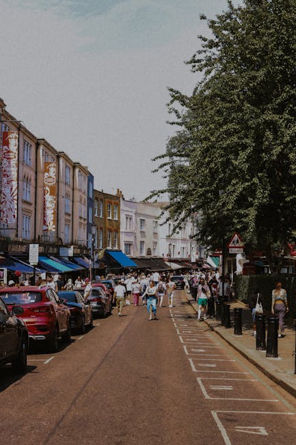A wide street scene in Notting Hill Gate during daytime, with a concrete pavement on the right side and a row of parked cars lining the curb. On the left, there are multicoloured Victorian-style terraced buildings with commercial storefronts at ground level, some featuring awnings. Pedestrians, including individuals carrying boxes and moving supplies, are walking along the pavement and across the street, suggesting a home relocation or moving process. Overhead, mature trees provide partial shade, and the sky is clear with bright natural light illuminating the scene. In the background, a blue van is parked, likely used for furniture transport or packing and moving activities, with some people seen loading or unloading items. The overall atmosphere reflects a busy urban environment appropriate for house removals and professional moving services, such as those offered by Man and Van Holland Park, supporting efficient moving logistics in central London neighborhoods.