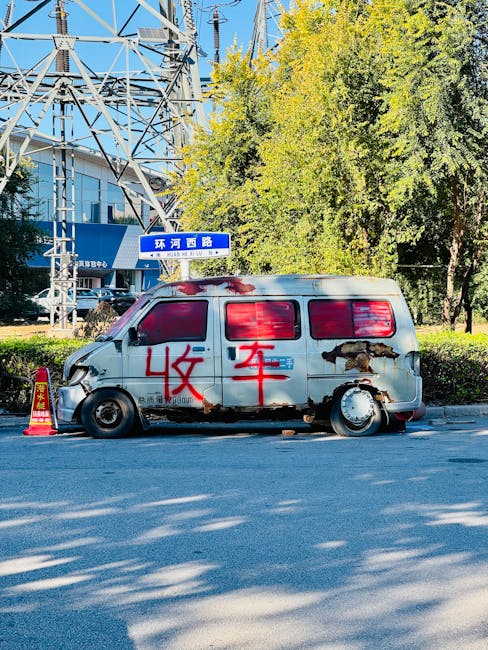 A weathered, silver delivery van parked on a city street during daylight hours, with visible rust patches and damage on its side, including a large hole near the rear wheel. The van's windows are covered with red spray-painted markings, and large Chinese characters are painted in red on the side. The vehicle is stationary, and the scene includes a traffic cone placed near the front left corner of the van. In the background, there are tall electrical transmission towers, a blue building with signage, and lush green trees under a clear, blue sky. The setting suggests an urban environment, possibly involved in home relocation or furniture transport, with the van ready for loading or unloading as part of a house removal service. Man and Van Holland Park occasionally use such vehicles for efficient moving logistics, packing, and furniture transport services.